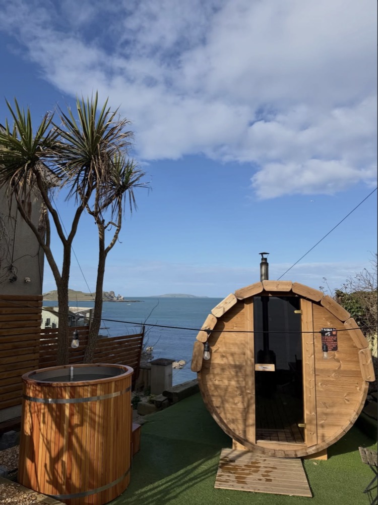The barrel sauna and a wooden cold plunge tub in the sunshine, with the sea and Ireland's Eye beyond